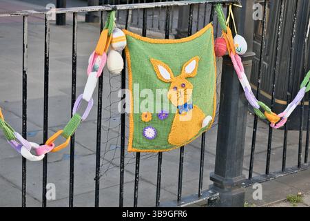 Knitted Easter decorations hang from a black metal fence, including a bunny bag, knitted eggs and a colorful knitted chain in Ripon, Yorkshire, UK. Stock Photo