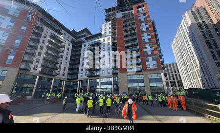 Wide shot of a large group of construction workers in front of apartment buildings on a sunny day, wearing high-visibility clothing and hard hats in L Stock Photo