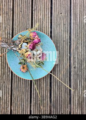 Dried blossoms of red roses and various flowers create a colorful floral arrangement on a light blue plate against a wooden background Stock Photo