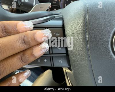 England UK, 27.04.2025. Womans decorated finger nails using hand controls alongside the steering wheel on a modern car. Stock Photo