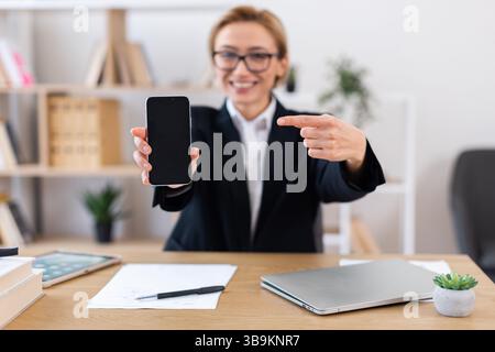 Businesswoman pointing at smartphone screen while sitting at desk with equipment Stock Photo