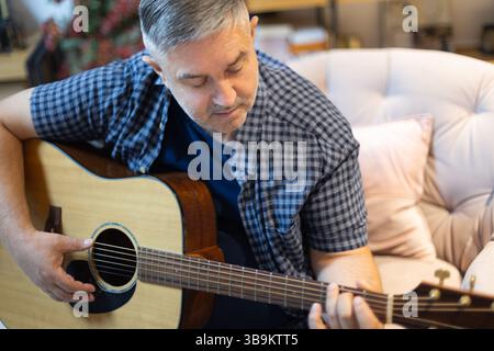 Portrait of a mature man sitting in a cozy chair at home, focused on playing an acoustic guitar. A peaceful moment of hobby, music Stock Photo