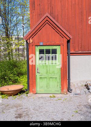 A vibrant green door with a triangular roof, set against a rustic red wooden building. The door features a glass panel and is surrounded by greenery a Stock Photo