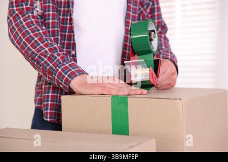 Man using dispenser with adhesive tape to seal cardboard box indoors, closeup Stock Photo