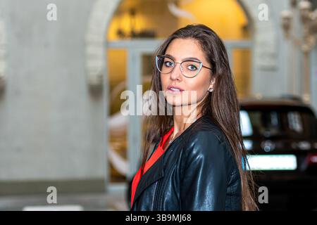 Confident woman with long hair wearing glasses stands in a stylish urban environment. Stock Photo