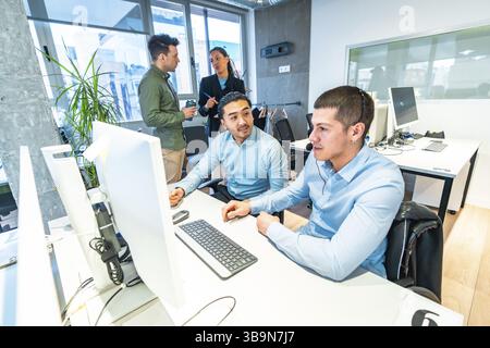 Two customer service representatives working on computer in a busy call center, assisted by their manager, with diverse colleagues standing in backgro Stock Photo