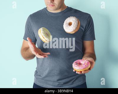 Juggler man juggles donuts on blue background. Doughnuts delivery or unhealthy diet concept. Flying glazed donuts with sprinkles and desiccated coconu Stock Photo