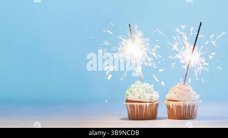 Two cupcakes decorated with colorful sprinkles and sparklers. Festive cupcakes on blue background with copy space left for text or design. Banner Stock Photo