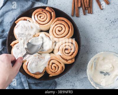 Woman's Hand Spreading Frosting on Cinnamon Rolls in Skillet. Vegan swedish cinnamon buns Kanelbullar with pumpkin spice, topping vegan cream cheese. Stock Photo