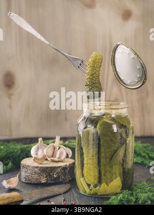 The fork holds a pickled cucumber that just has been taken from glass jar. Pickled cucumbers on wooden background. Pickles in glass jar with garlic, p Stock Photo