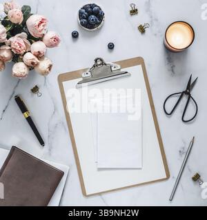 Wedding mock up. Flat lay with blank greeting card, envelope. Clipboard, flowers, scented candle on white marble. Feminine home office mock up with co Stock Photo