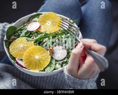 Woman in jeans at bed, holding vegan salad bowl with oranges, spinach, arugula, radish, nut.Top view.Vegan breakfast, vegetarian food, diet concept.Gi Stock Photo