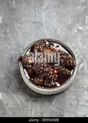 Delicious jam with baby pine cones in small bowl. Traditional Siberian dessert - young pine cones jam on gray textured background. Top view or flat la Stock Photo