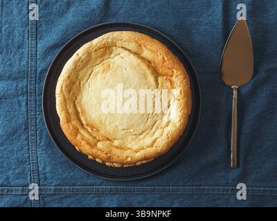 Plate with whole cheesecake pie on table with jeans tablecloth. Classic cheesecake flat lay or top view. Vertical Stock Photo