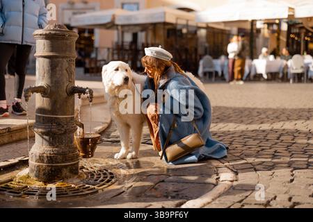A tender moment at a Roman fountain as a woman crouches beside her fluffy white dog, sharing eye contact and care during a walk through a lively piazza Stock Photo