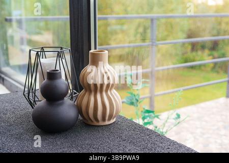 Two ceramic decorative vases or candlesticks in different shapes and colors are displayed on sleek table. Beautiful green landscape is visible through Stock Photo