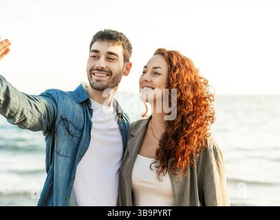 Smiling man gestures while standing next to his partner on a beach, both looking ahead and sharing a cheerful, relaxed moment by the ocean. Lifestyle. Stock Photo