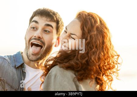 Young couple laughing and making silly faces together during sunset by the ocean, capturing a carefree and joyful moment in their relationship. Lifest Stock Photo