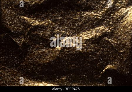 Golden foil with dark, glossy texture creates a dramatic, reflective surface. Crumpled, burnished folds add moody elegance, while shimmer and glare en Stock Photo