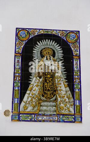 Ceramic tile mural of “Ntra. Sra. del Carmen” on a white wall in Seville, Spain, showing traditional Catholic devotional art with ornate border. Stock Photo