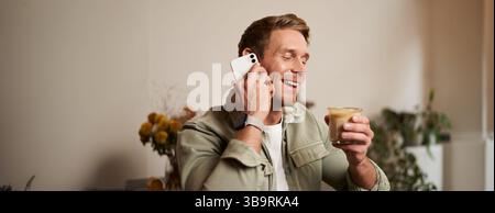 Portrait of young handsome man talking on the phone, drinks coffee and laughs. A guy sits in cafe having a nice, pleasant conversation over cup of Stock Photo