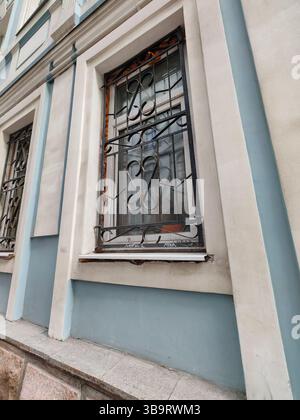 A close-up of a barred window with decorative ironwork, set in a pale blue and white facade. Stock Photo