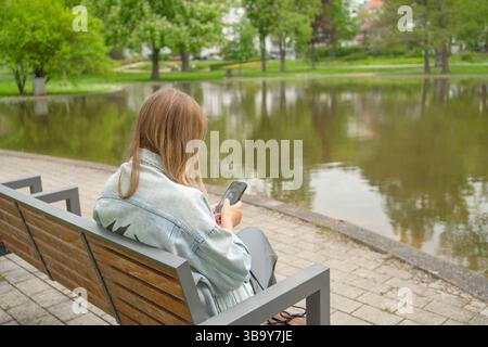 A woman sits on a park bench by a tranquil lake, focused on her smartphone. Surrounded by lush greenery, she enjoys a peaceful moment in nature, trees Stock Photo
