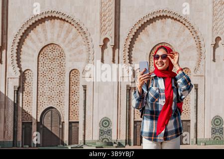 Smiling tourist wearing red hijab and sunglasses using smartphone in front of Hassan II Mosque in Casablanca, Morocco Stock Photo