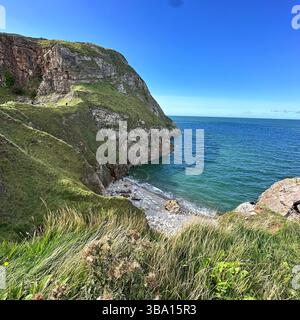 View over Angel Bay (Porth Dyniewaid) on the Little Orme, North Wales, with calm sea, blue sky, and lush green cliffs on a clear day Stock Photo