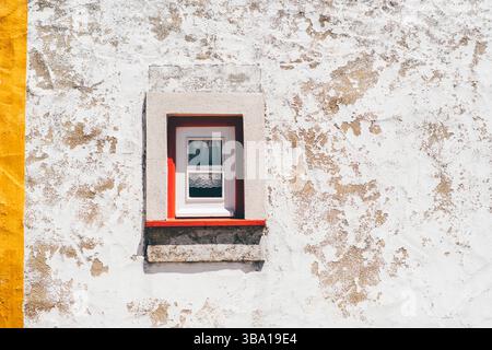 Close-up of a small white-framed window with lace curtain, set in a textured white, beige stucco wall with a yellow stripe: rustic Mediterranean charm Stock Photo