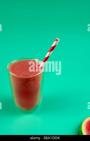 A glass of watermelon juice with a straw in on a green table. The juice is a bright red color and the straw is red and white. Stock Photo