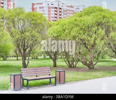 Modern park scene featuring wooden bench between two trash bins on paved path, surrounded by lush green trees and residential buildings in background. Stock Photo
