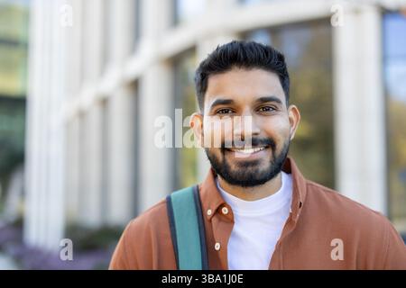 A smiling young Indian man with a backpack stands outdoors, looking at the camera. He is wearing a shirt and smiling. Stock Photo