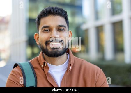 Smiling Indian man poses confidently in front of a modern building, possibly a student or professional with a backpack. Stock Photo