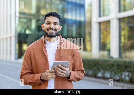 A smiling man stands outdoors, holding a tablet. He wears a casual orange shirt and a white t-shirt, with a modern building in the background. Stock Photo
