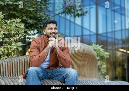 A cheerful man sits on a modern bench in front of a building, smiling and looking to the side. Stock Photo