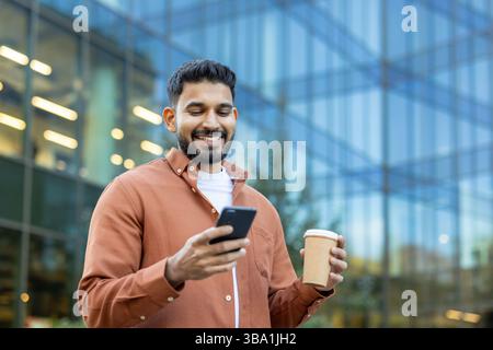 A smiling man is using his phone outdoors. He holds a coffee cup, showing a relaxed and happy demeanor near an office building. Stock Photo