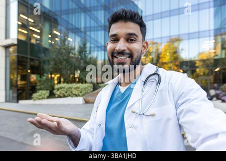 A cheerful doctor gestures towards the viewer, inviting or presenting, in front of a modern glass building. Stock Photo