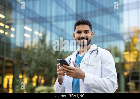 A smiling doctor looks at the camera while holding his smartphone outdoors, near a modern office building. Stock Photo