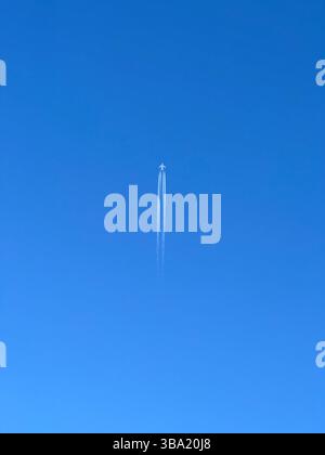 An airplane streaks across a bright blue sky, leaving a wispy contrail in its wake. Stock Photo