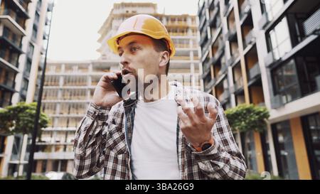 Angry male architect in orange hard hat talks swears on phone at construction site. Builder in plaid shirt emotionally displeased negotiates by cell p Stock Photo