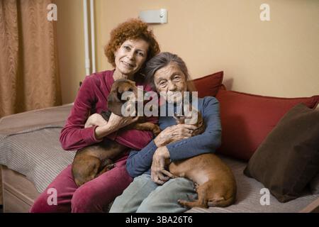 Elder woman and her adult daughter together with two dachshund dogs on sofa indoors spend time happily, portrait. Theme of mother and daughter relatio Stock Photo