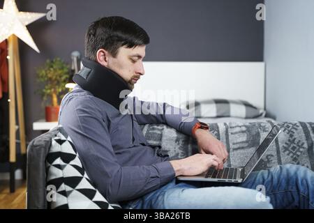 Man employee after accident working at home behind laptop and feels pain in neck, uses black neck collar support. Male with neck injury freelance prog Stock Photo
