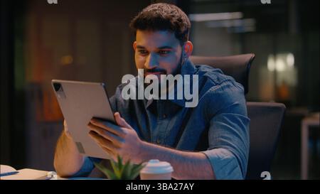 Smiling Indian Arabian man satisfied businessman ethnic male guy business entrepreneur in dark office late night working using digital tablet gadget Stock Photo