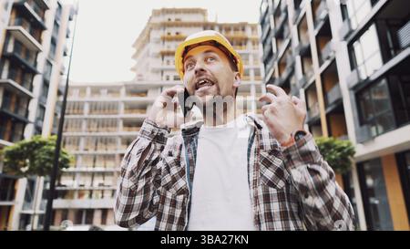 Angry builder, dissatisfied with deadline of work, swears at stress while talking to foreman on phone background of construction site. Wrathful archit Stock Photo