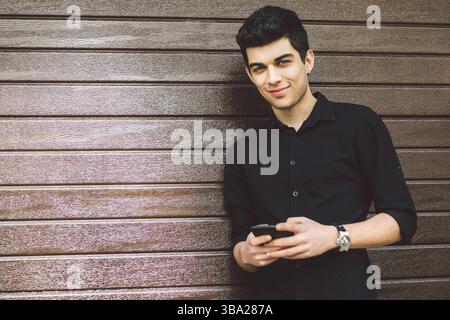 Portrait of young handsome Turkish male model. Mediterranean race brunette in black shirt is using a hand phone of a technology businessman on the str Stock Photo