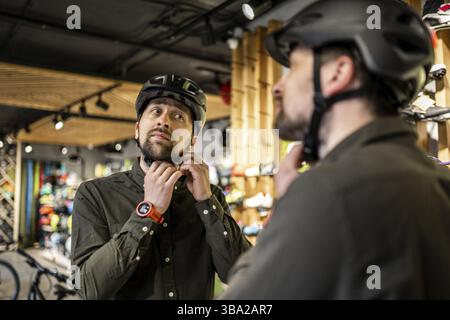 Caucasian man trying on black bicycle helmet near mirror in sporting goods store. Male buyer chooses safety helmet for cycling. Shopping in bicycle st Stock Photo