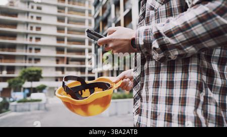 Young Engineer using smartphone on construction site. Architect using phone. Manager development building texting on smart phone. Builder makes notes Stock Photo