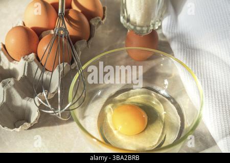 Broken egg in a glass bowl ready for whipping. Fresh chicken eggs in a cardboard tray, whisk and white towel on a kitchen table Stock Photo