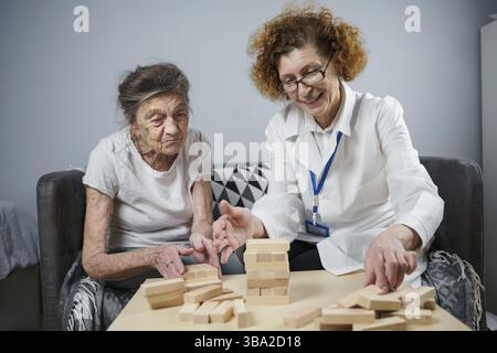 Senior woman practice skills, build wooden blocks, build tower and try not to let it fall, Jenga game. Old patient pull out block, place on top, suppo Stock Photo